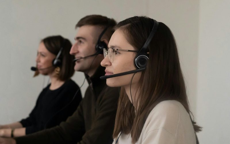 Side view of young positive operators in casual outfit with headphones with microphone in light workplace